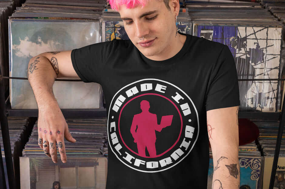 Man wearing "Made In California" Ghetto Rags T-shirt with retro design, standing in a record store with colorful hair and tattoos.