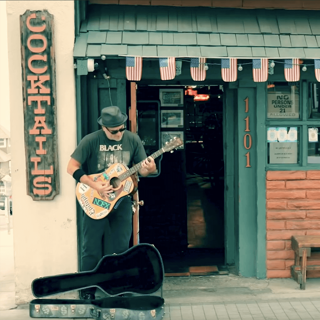 Musician playing guitar outside a bar with "Cocktails" sign, wearing a hat and sunglasses, with an open guitar case on the ground.