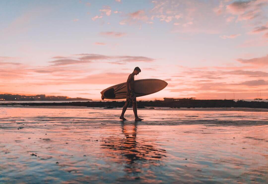 Surfer walking on beach at sunset carrying surfboard, reflecting serene ocean vibes and coastal lifestyle.