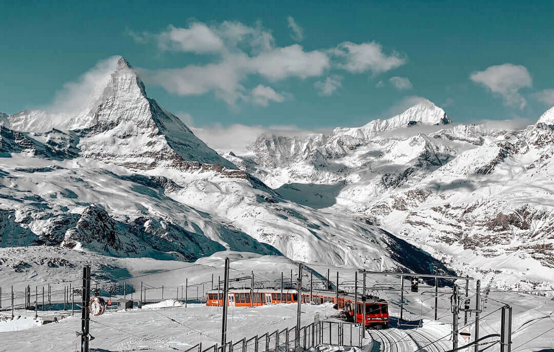 Mountain landscape with snowy peaks and a red train traveling through the winter scenery.