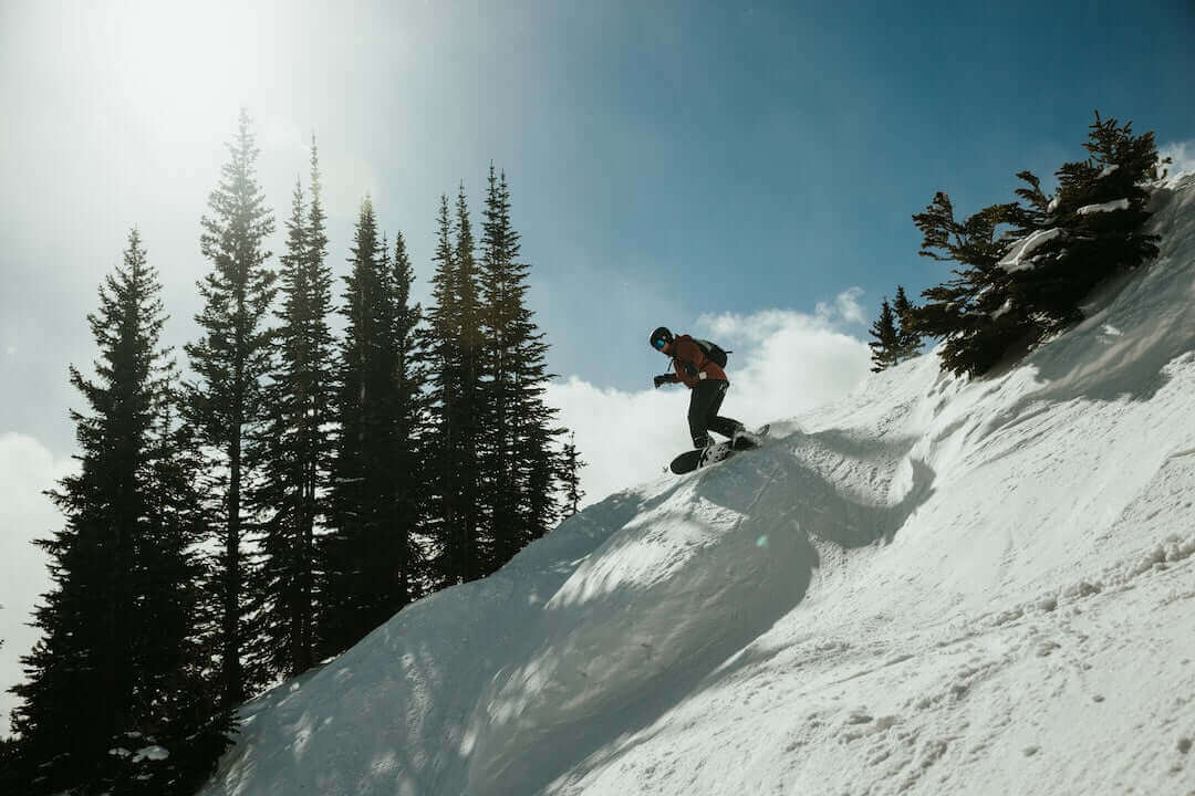 Snowboarder tackling a steep slope amidst sunlit trees and clear blue sky