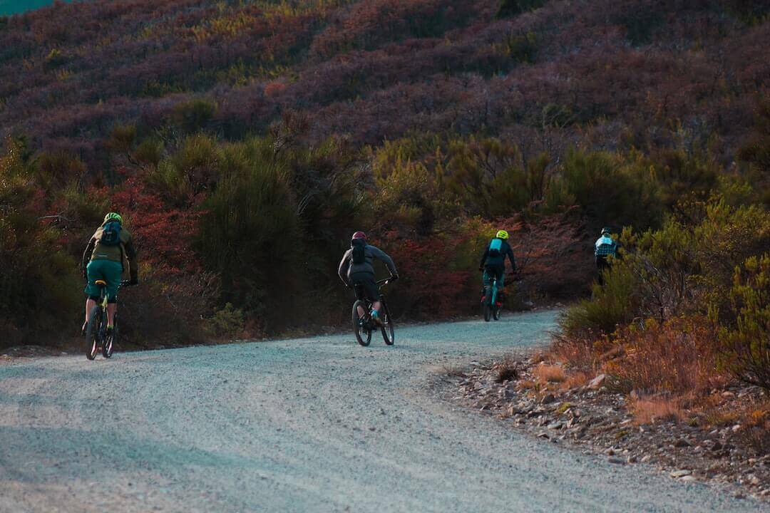 Dirt bikers wearing streetwear riding on a winding gravel trail amidst lush greenery and rugged terrain.