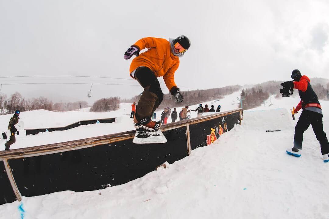 Snowboarder in orange jacket doing a trick on a snowy slope with people watching in the background.