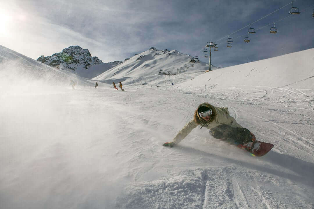 Snowboarder carving down a snowy mountain slope under a clear sky with chairlifts in the background.