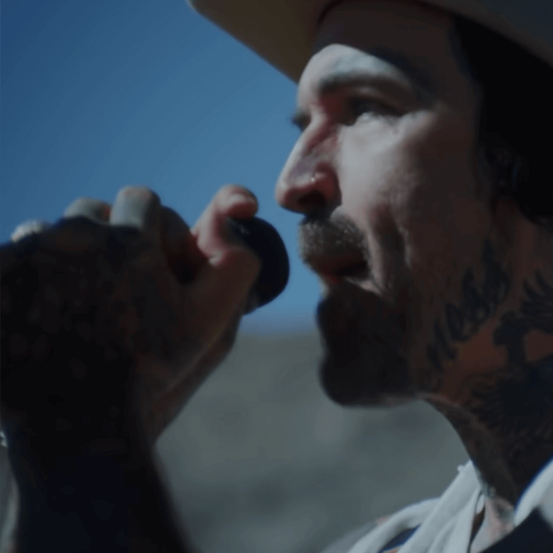 Music artist holding microphone and singing outdoors, showcasing tattoos and wearing a hat.