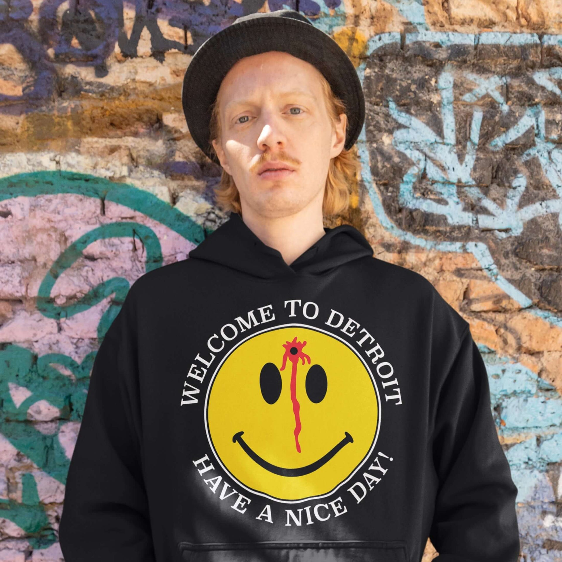 Person wearing Welcome to Detroit Hoodie with smiley face graphic against graffiti wall